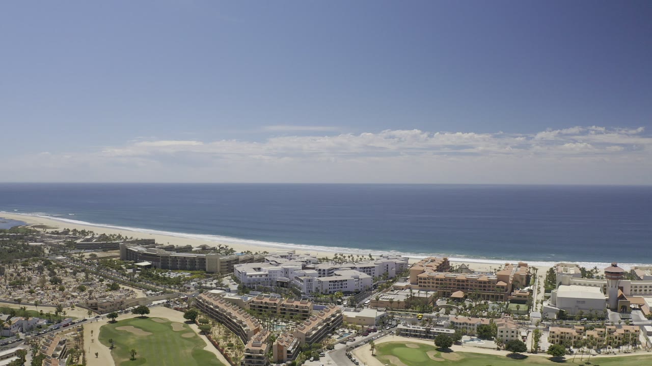High aerial frame over Los Cabos, Mexico, showing beachfront buildings, roads, a green course area, pale sand, blue ocean, and no visible people.
