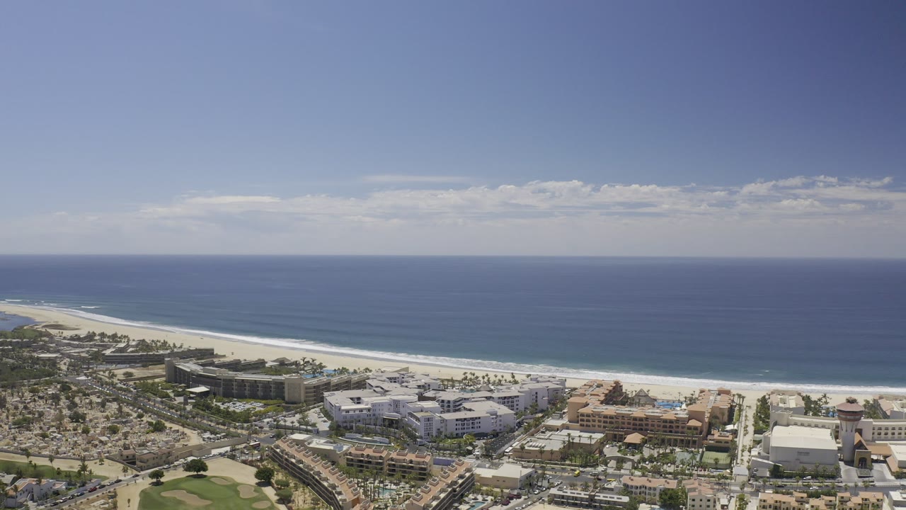 Wide aerial frame over Los Cabos, Mexico showing city blocks, beachfront buildings, a pale sand shoreline, blue ocean, and no visible people.