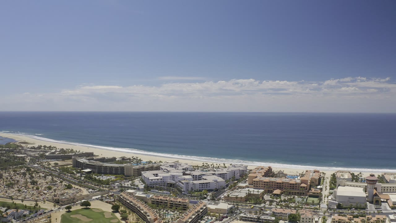 Aerial view of Los Cabos, Mexico with the beach and surf line stretching left to right, large beachfront structures below, and no visible people.