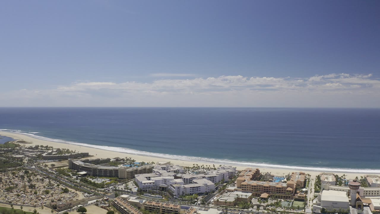 Opening aerial frame over Los Cabos, Mexico, with blue ocean, a pale beach, coastal buildings in the foreground, and no visible people.