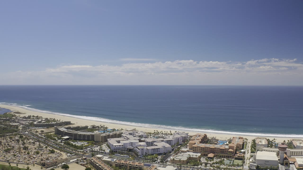 A wide Los Cabos aerial view shows the ocean horizon, bright beach strip, and clustered coastal buildings with no visible people.
