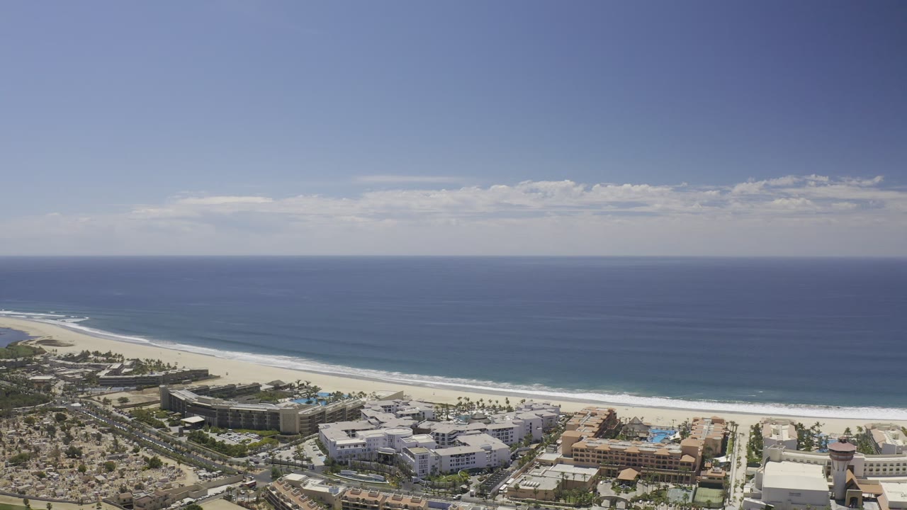 Aerial Los Cabos coastline view with the ocean horizon across the background, pale beach below, resort blocks near shore, and no visible people.
