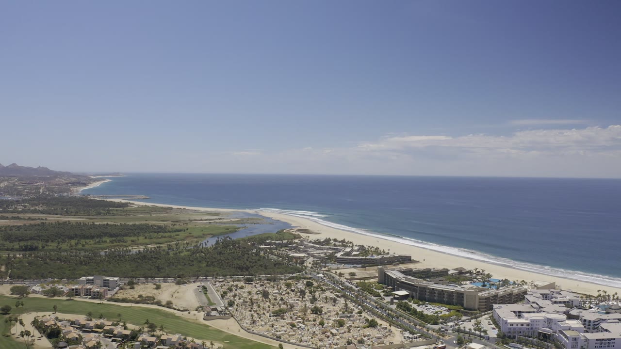 Aerial Los Cabos coastline frame with the ocean horizon high in the image, a bright beach curving along the shore, green terrain inland, and no visible people.