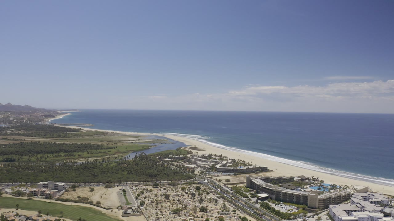 Aerial view of beach, blue ocean, resort buildings, palms, and distant hills.