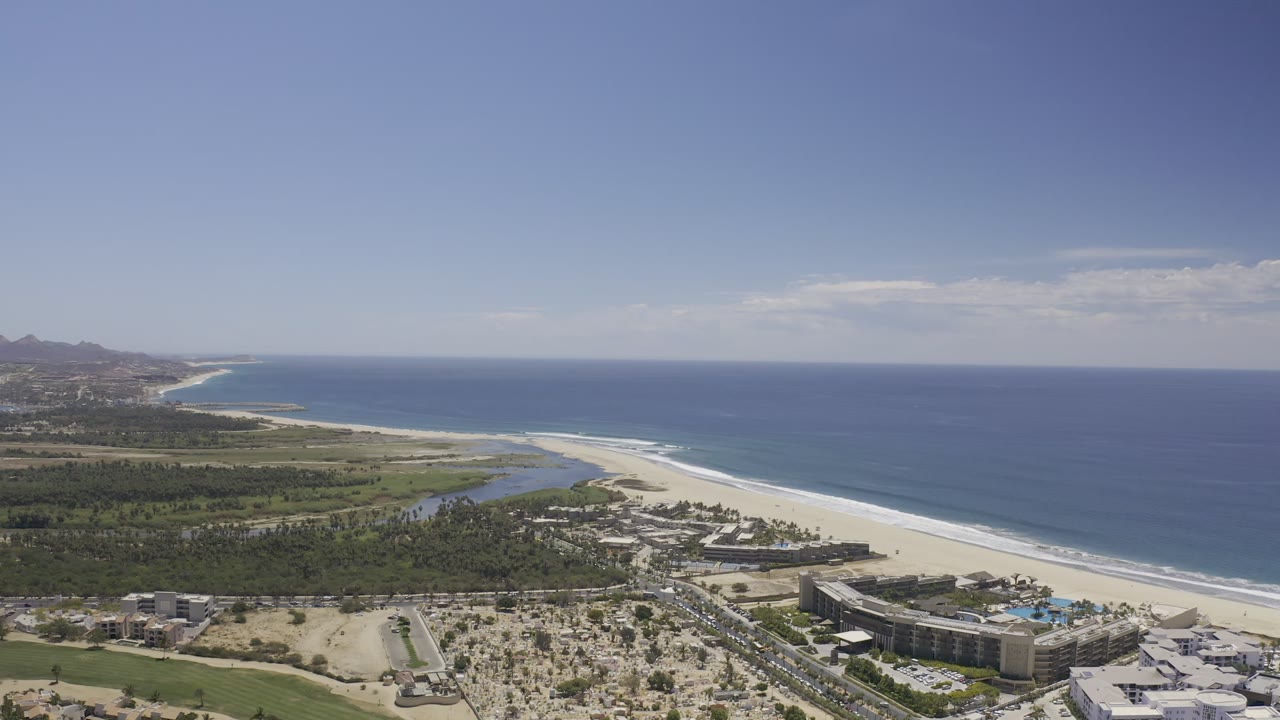 The Los Cabos aerial frame looks along the shoreline with deep blue water on the right, pale sand below, a lagoon or river behind the beach, resort buildings, and no visible people.