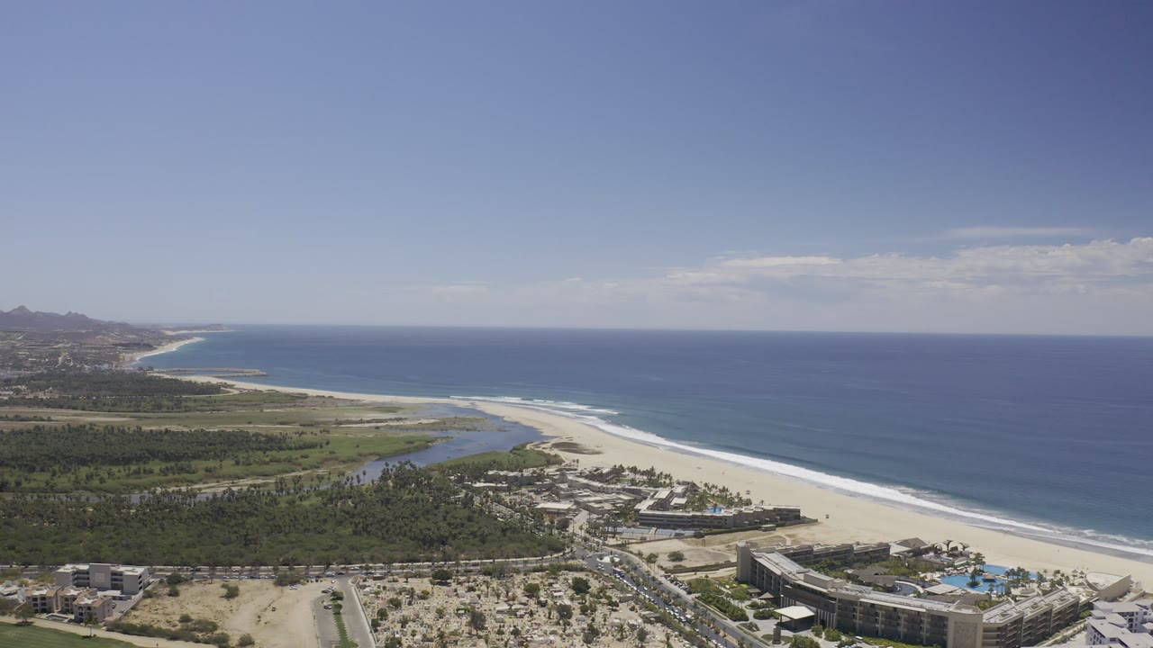 Aerial view of a curving beach, blue sea, lagoon, palms, and coastal buildings.