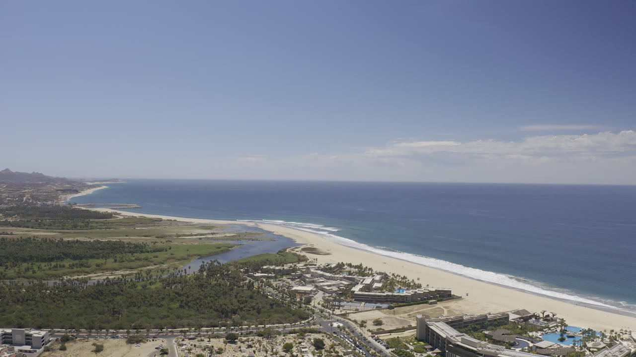 Aerial view of Los Cabos with the ocean filling the right side, a long beach below, a lagoon behind the sand, resort rooftops near the bottom edge, and no visible people.
