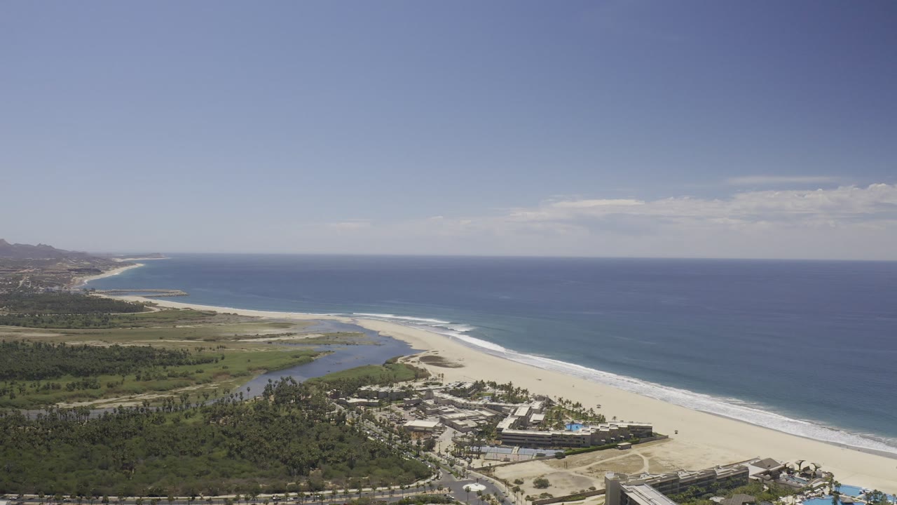 Wide aerial frame over the Los Cabos coastline with blue ocean at right, pale beach through the center, inland lagoon at left, beachfront buildings below, and no visible people.