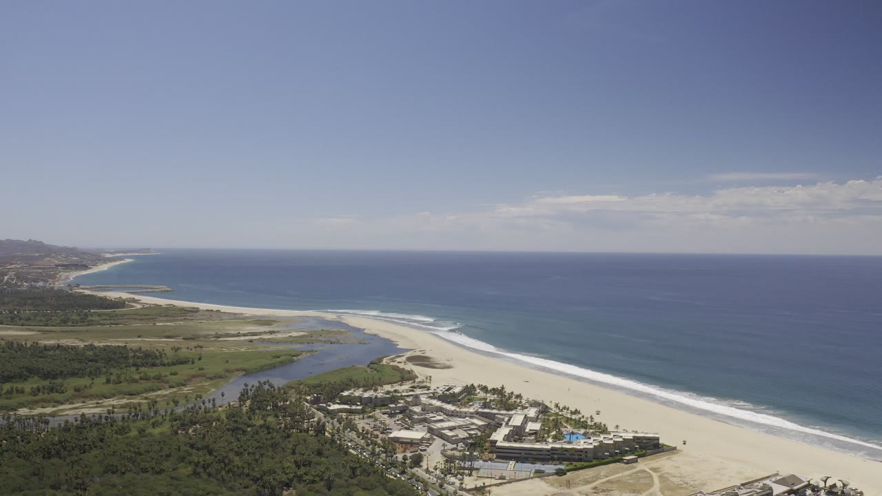 Opening aerial frame over Los Cabos, Mexico, showing resort buildings at the lower edge, a pale beach, a lagoon mouth, blue ocean, distant coast, and no visible people.