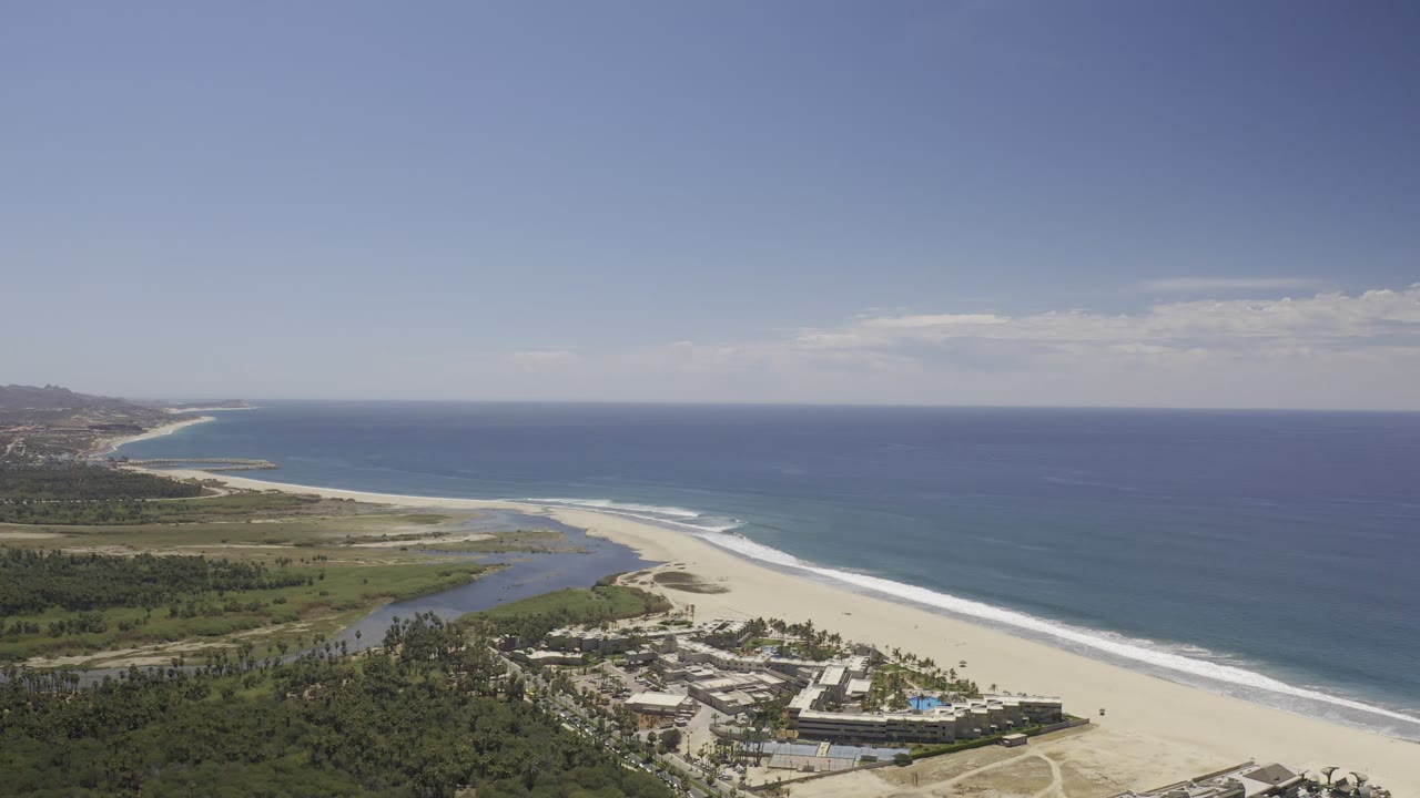 Aerial frame in Los Cabos, Mexico, with the resort slightly more centered below, the sandy beach stretching diagonally, ocean water to the right, and no visible people.