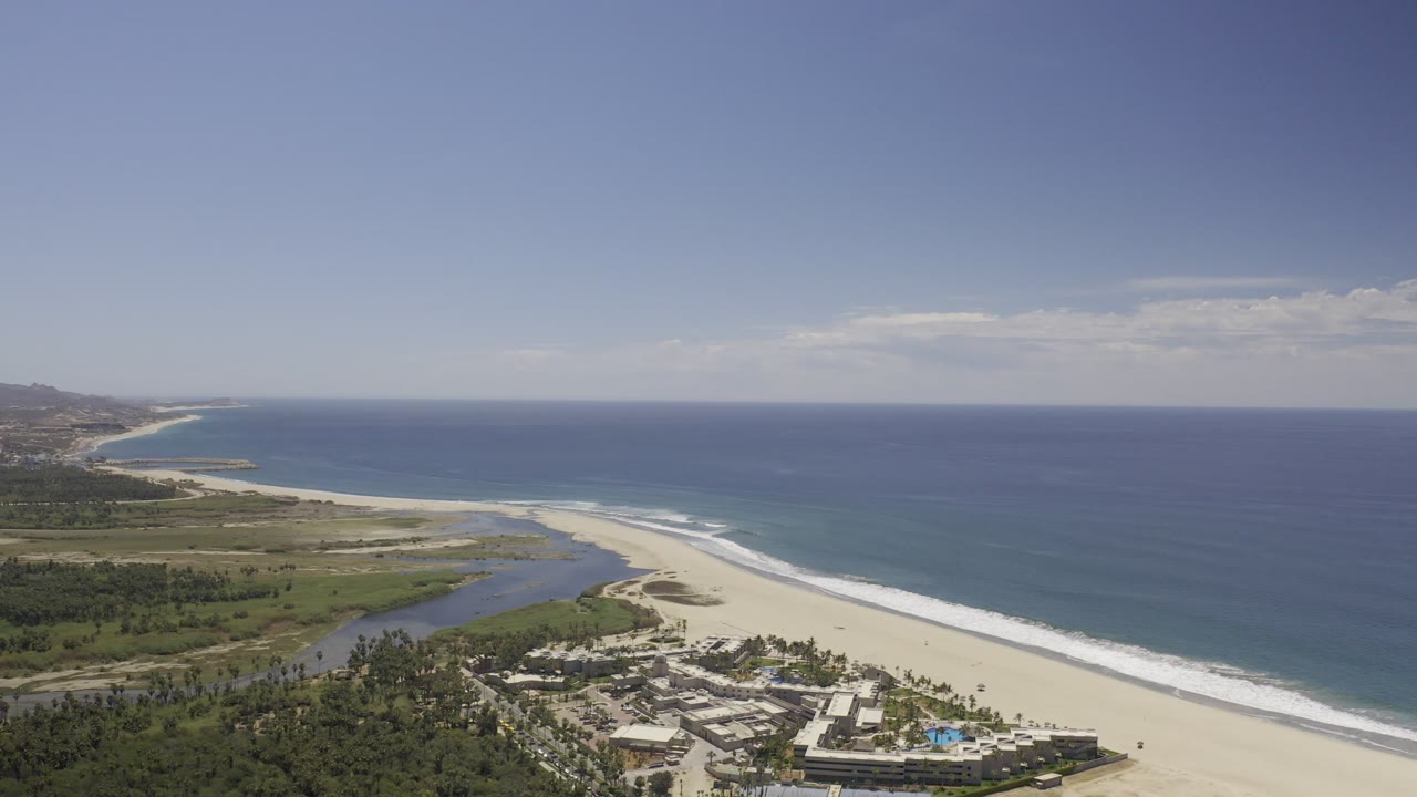 Opening aerial frame over Los Cabos, Mexico, with blue ocean, a long sandy beach, a lagoon, palm-covered grounds, hotel buildings, and no visible people.