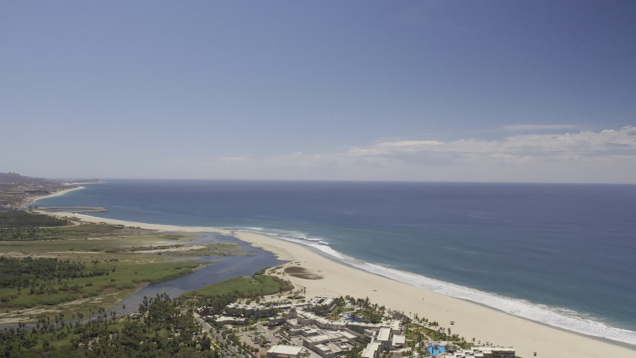 Aerial view over Los Cabos, Mexico, with a pale sand beach, blue ocean, lagoon water, green shoreline, low buildings at the bottom edge, and no visible people.