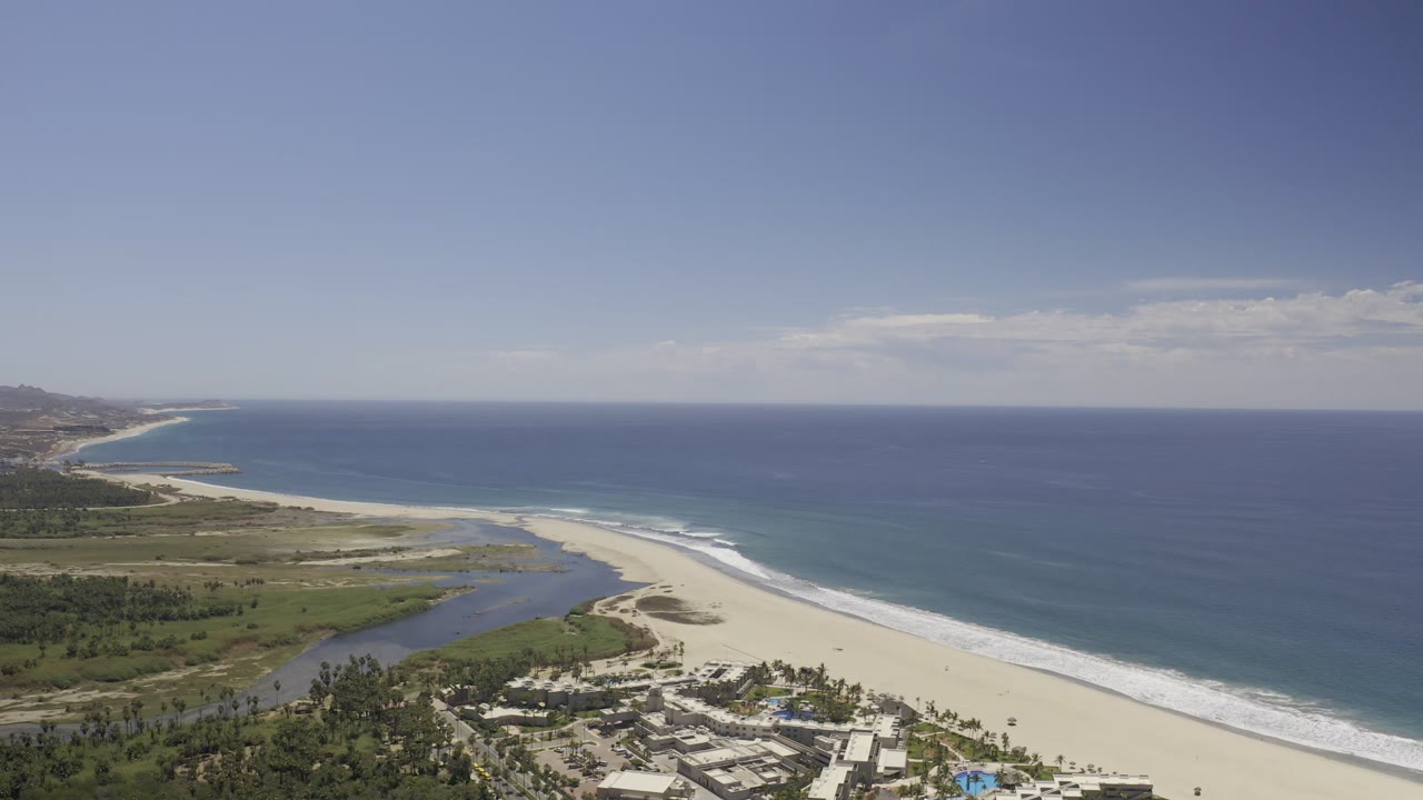 Los Cabos aerial coastline frame with the ocean horizon high in view, a curving beach and surf line through the center, lagoon water inland, and no visible people.