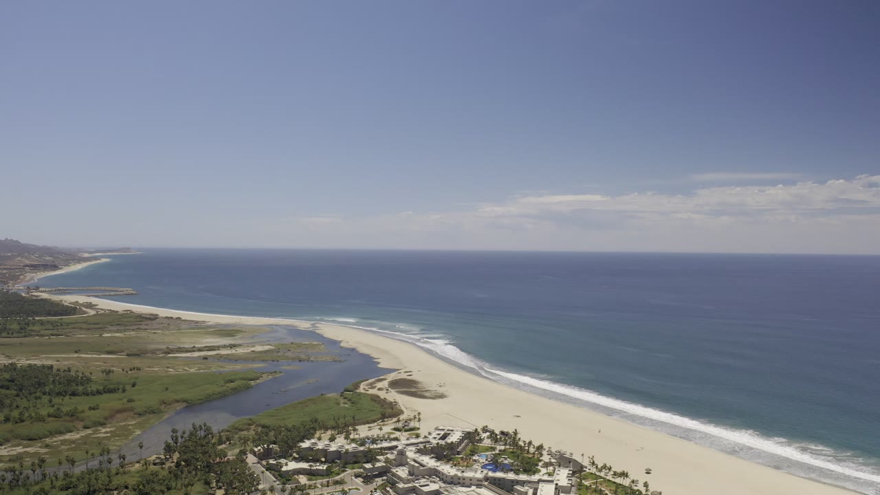 High aerial frame of the Los Cabos coast showing a long surf line, pale sandbar, inland water channel, low greenery, distant hills, and no visible people.