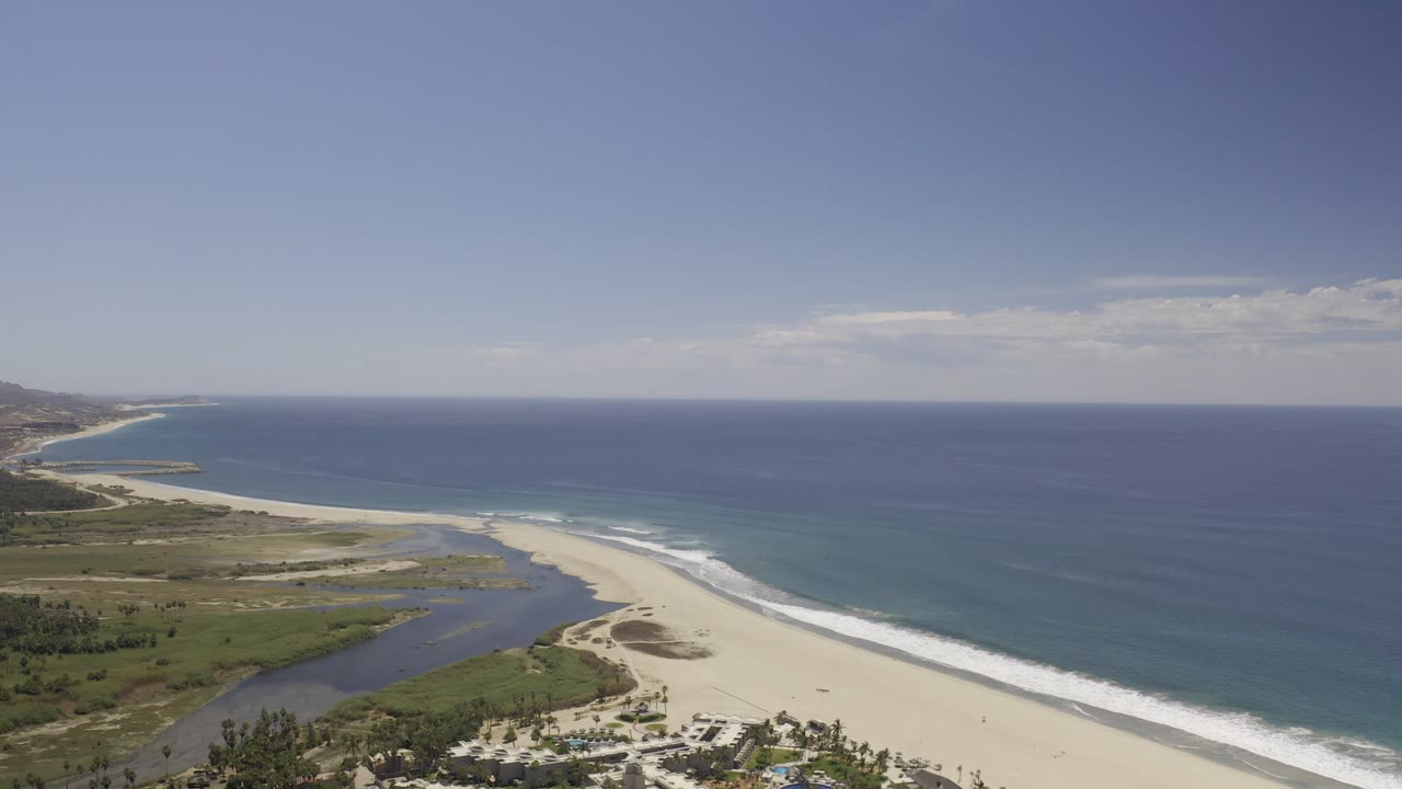 Elevated view over a Los Cabos beach where white waves meet pale sand, a lagoon cuts inland, and no visible people appear in the frame.
