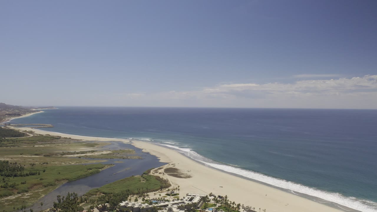 Aerial coastline with pale sand, white surf lines, lagoon, and open sea.