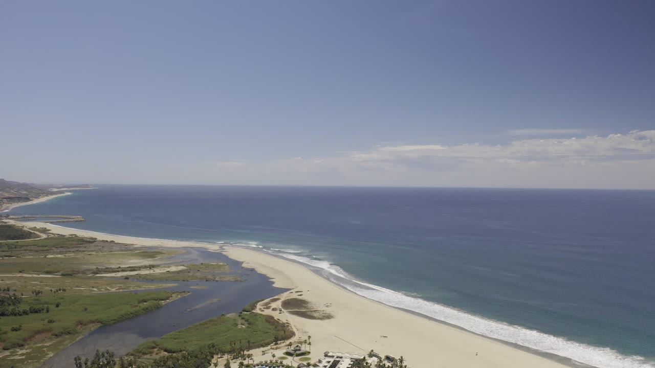Aerial view along the Los Cabos beach, with the lagoon slightly more centered, surf breaking in white bands, distant shoreline buildings, and no visible people.