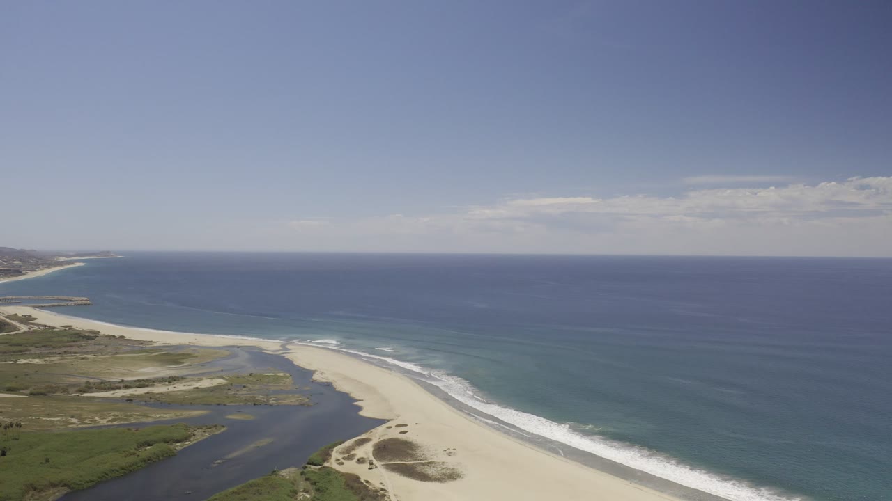 High aerial view over a Los Cabos beach where lagoon water meets pale sand beside turquoise surf, with no visible people.