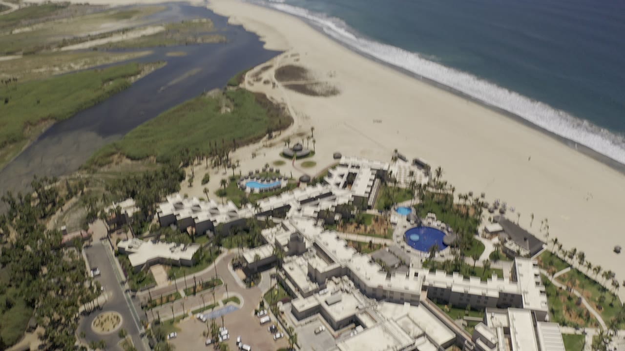 Aerial view of resort pools beside a wide beach and ocean surf.