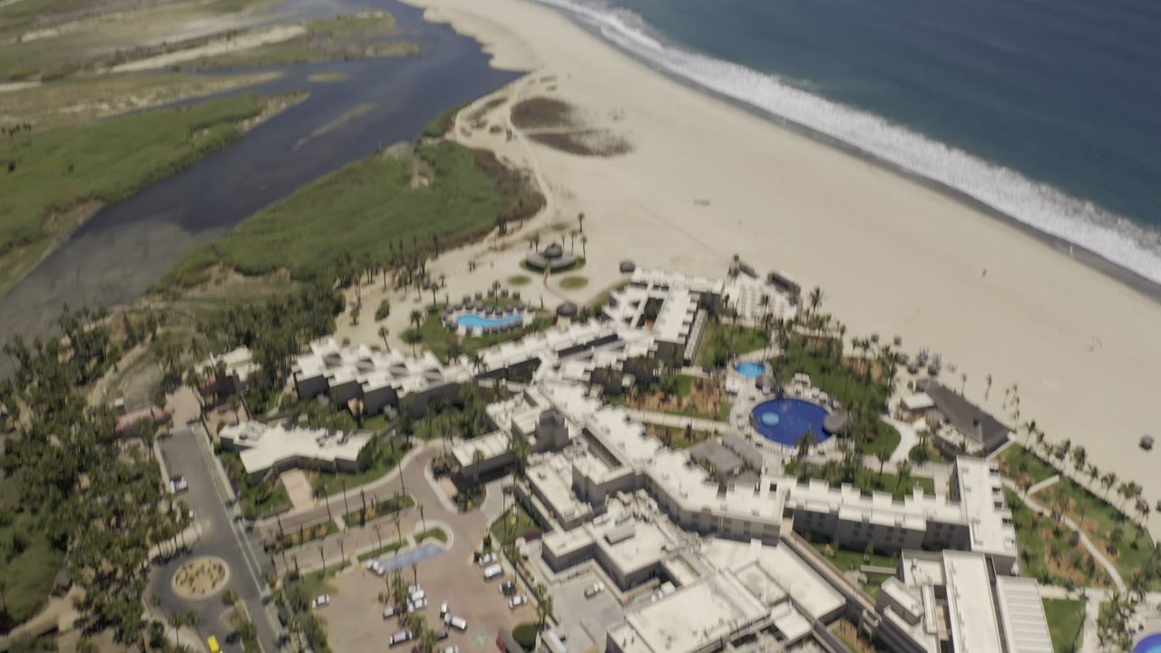 High drone view in Los Cabos, Mexico, with hotel grounds and circular blue pools in the lower frame, pale sand in the center, lagoon water at left, and ocean surf at right.