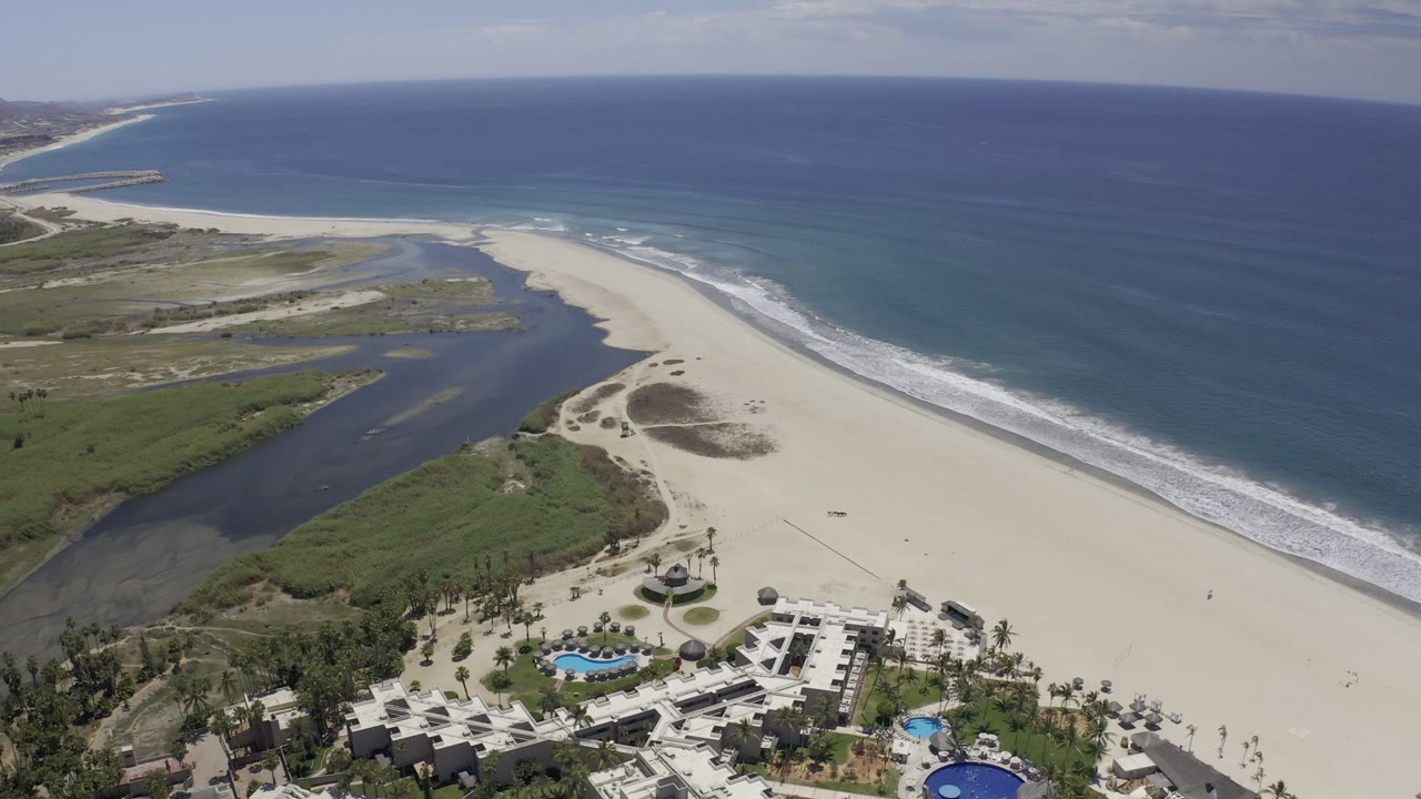 Aerial frame over Los Cabos, Mexico showing resort pools at the bottom, a long sandy beach, lagoon water to the left, blue ocean to the right, and no visible people.