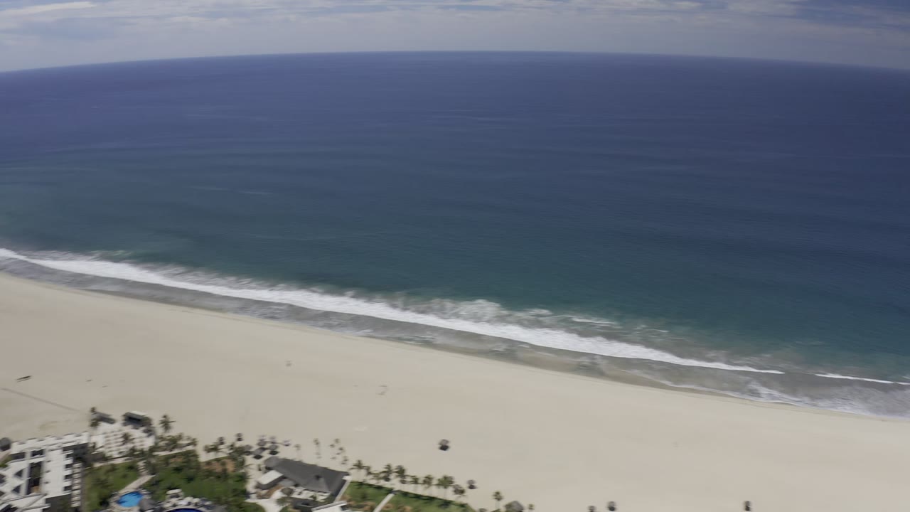 Aerial Los Cabos shoreline view with a long white surf band, open blue ocean, beach sand below, and no visible people.