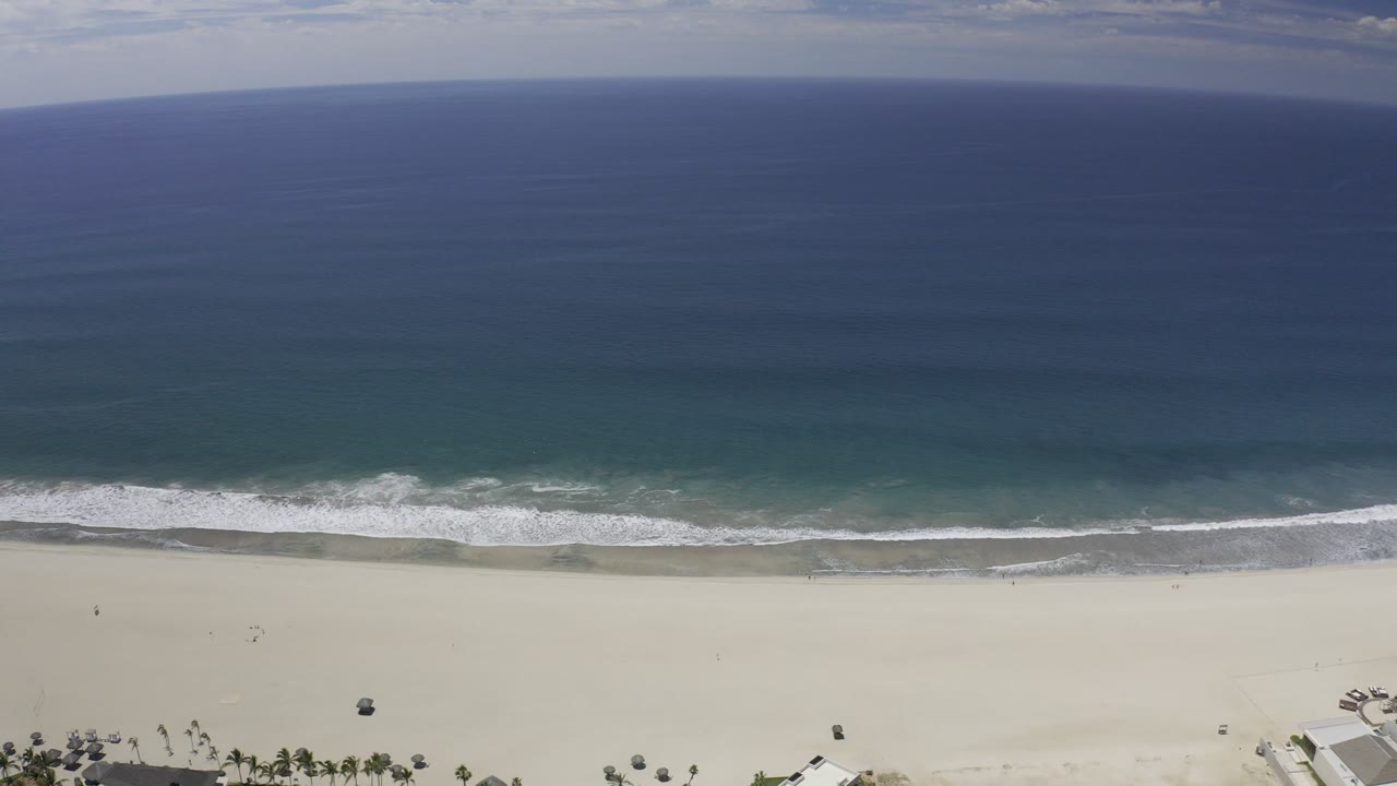Opening aerial frame over a Los Cabos beach, with deep blue water above, a white surf line at midframe, pale sand below, and no visible people.