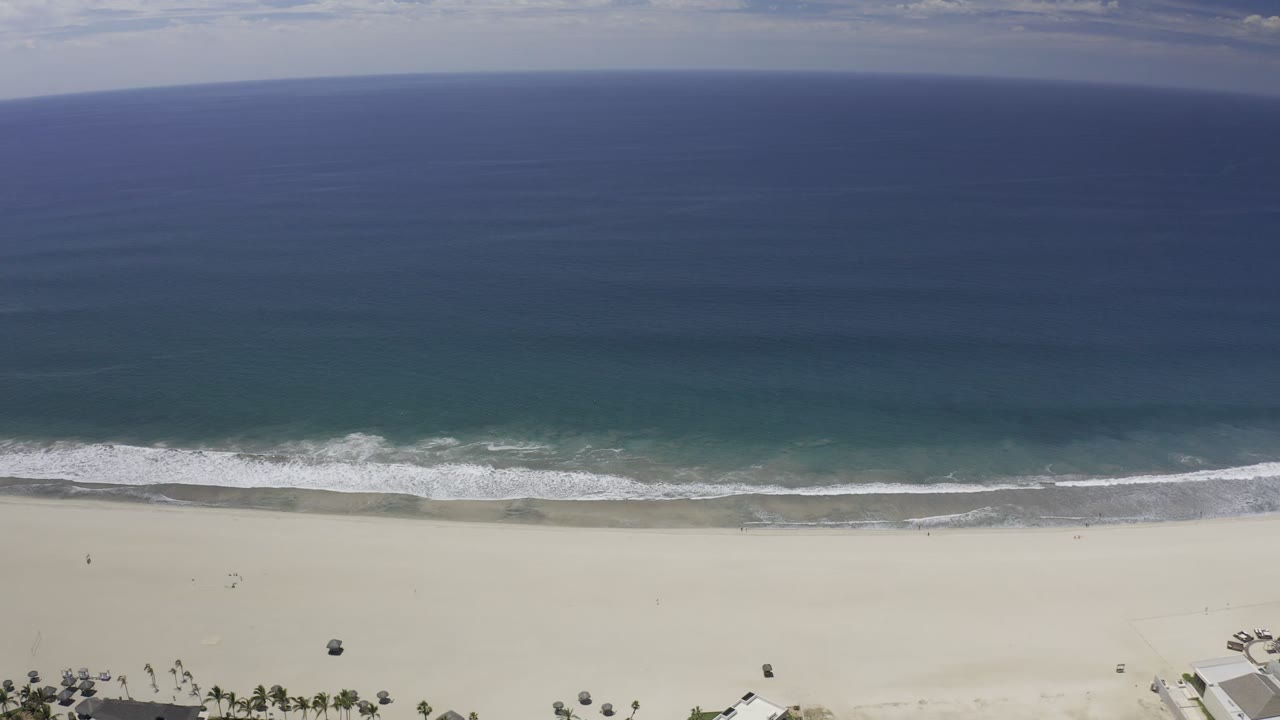 A second Los Cabos aerial frame shows the surf breaking slightly farther along the shoreline, with blue ocean, pale beach, and no visible people.