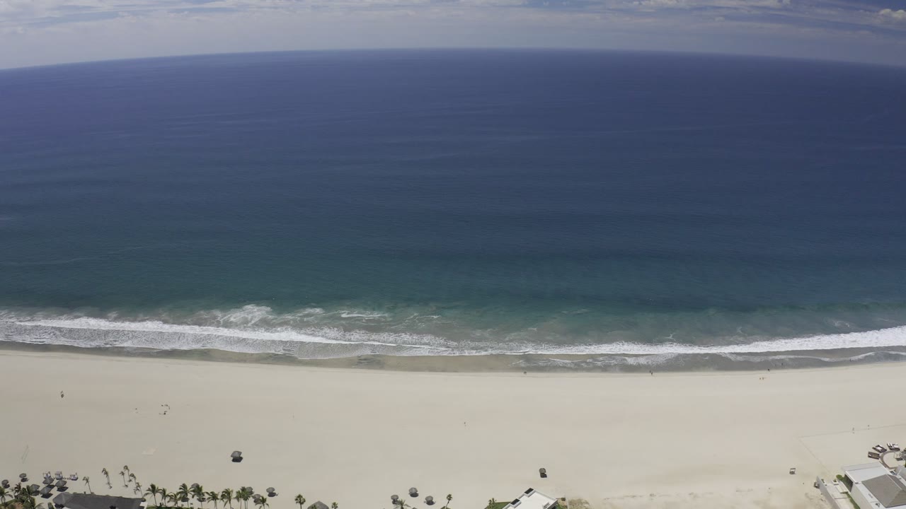 Opening aerial frame over the Los Cabos coastline, with deep blue ocean above a thin surf line, broad pale sand, palms, beachfront structures at the lower edge, and no visible people.