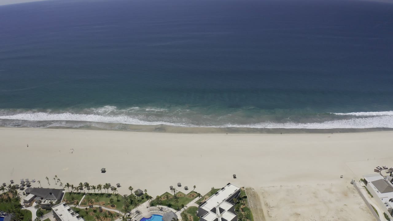 Aerial view of ocean surf, wide sand beach, and beachfront buildings.