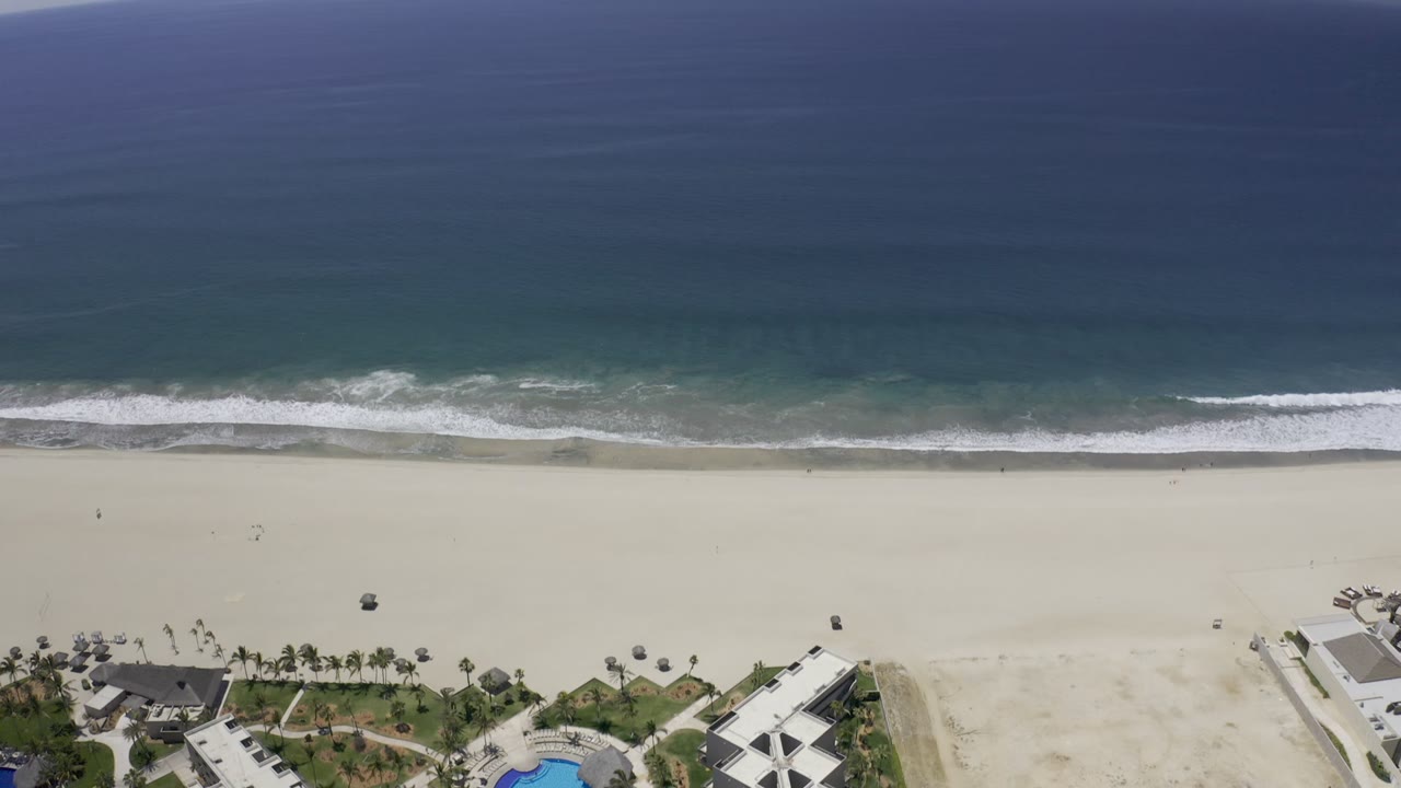 Aerial view in Los Cabos showing a broad band of ocean above white breaking waves, an open sandy beach, and a small resort or building edge below with no visible people.