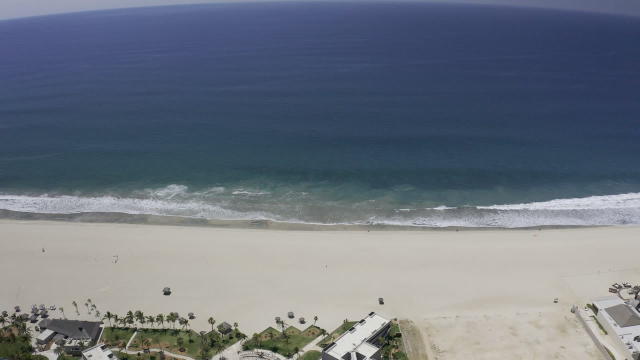 Aerial view of blue ocean, surf, pale beach, palms, and coastal buildings.