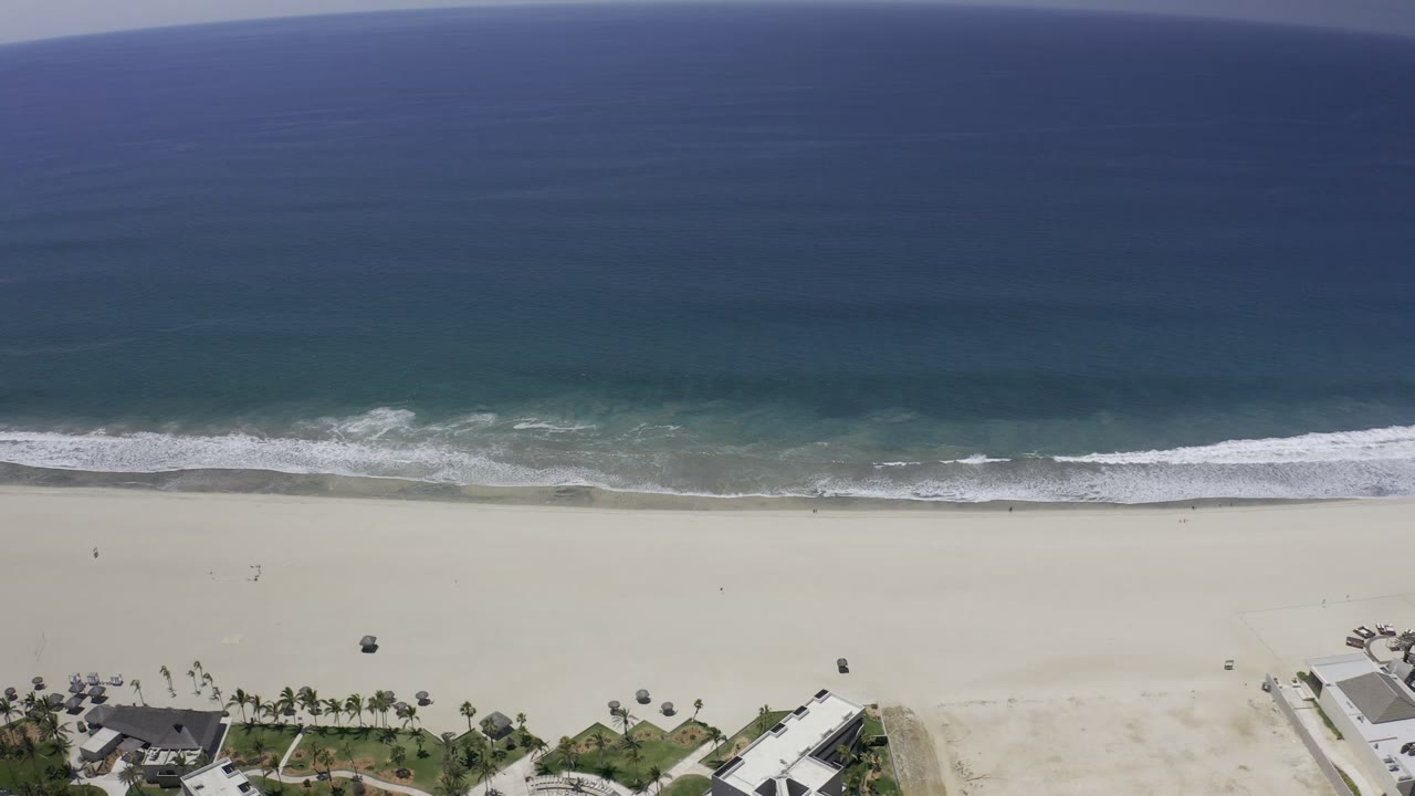 Aerial Los Cabos shoreline frame with the surf band slightly shifted across the sand, deep blue water above, beach below, palms and coastal buildings at the lower edge, and no visible people.
