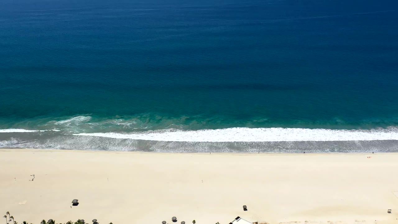 Los Cabos shoreline seen from above, with a long white wave line crossing the frame and broad light sand below it, with no visible people.