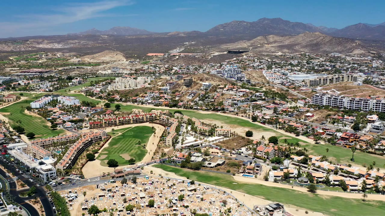 Opening frame of a wide Los Cabos, Mexico aerial showing golf course fairways, residential blocks, roads, dry hills, distant mountains, blue sky, and no visible people.