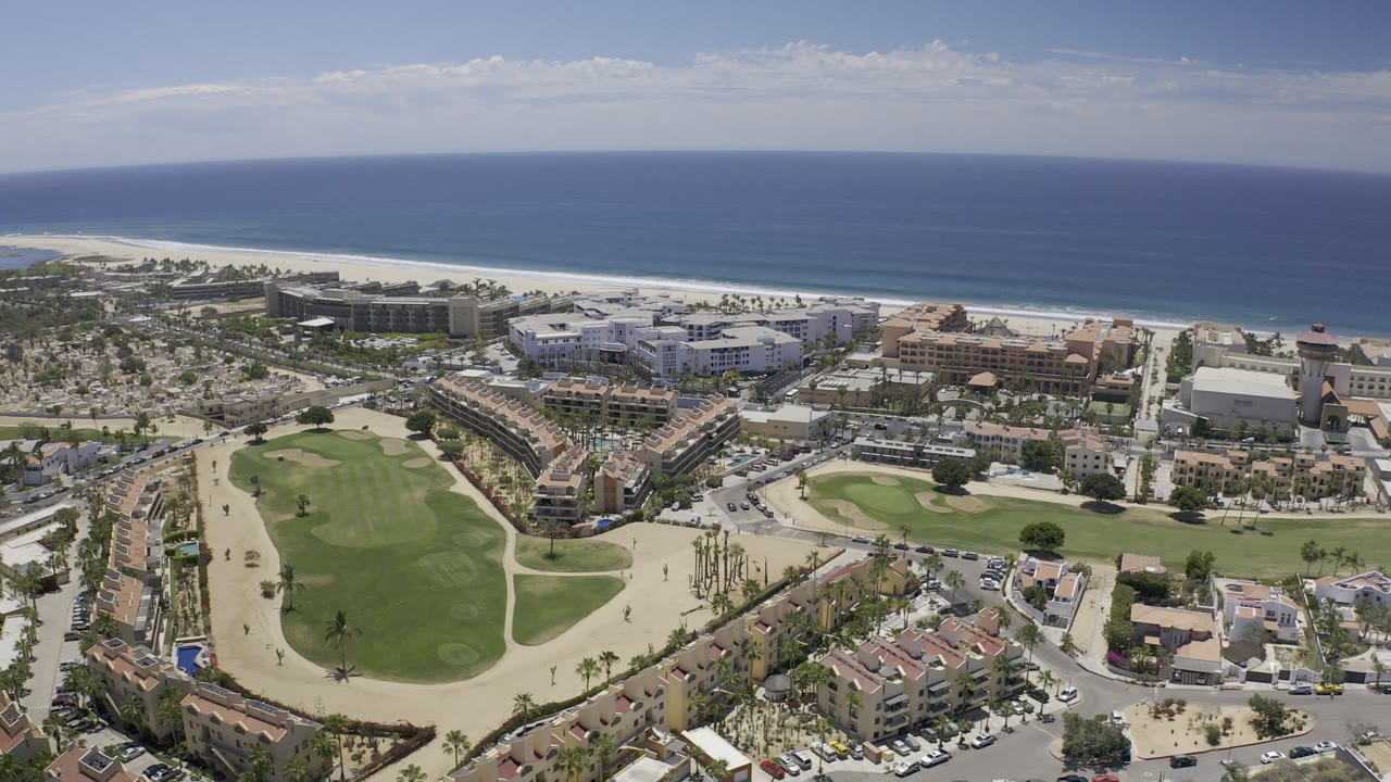 High aerial view over Los Cabos, Mexico, with golf fairways and roads in the foreground, resort buildings near the beach, blue sea beyond, and no visible people.