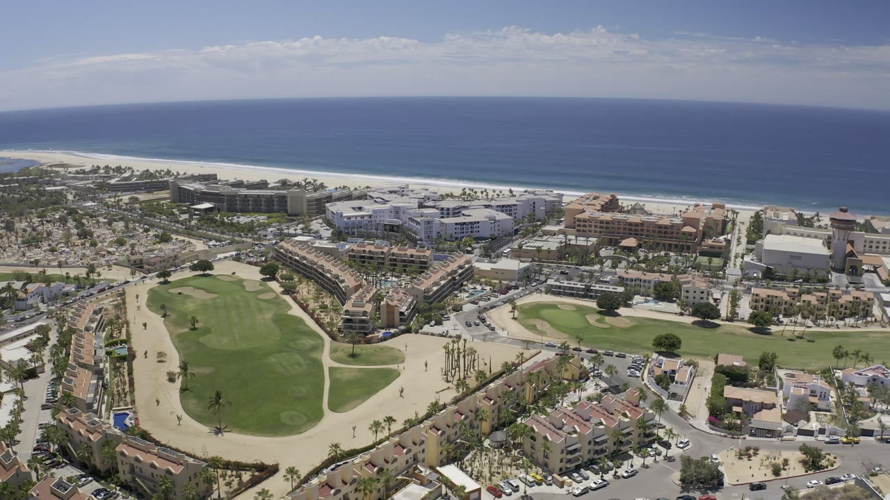 Aerial frame in Los Cabos, Mexico, showing green fairways below, a cluster of coastal buildings at center, the beach and ocean across the top, and no visible people.