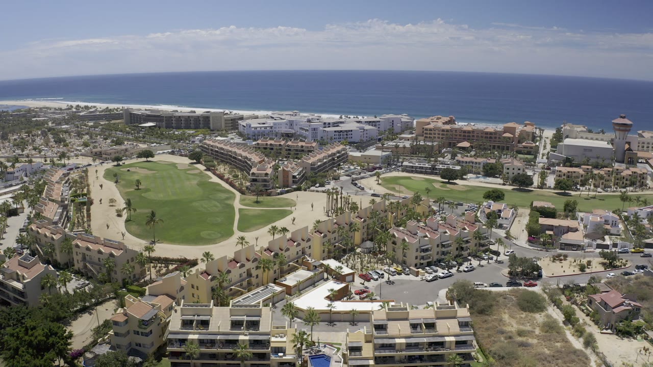 High aerial frame in Los Cabos, Mexico showing a rounded green lawn, clustered coastal buildings, a road with parked vehicles, palm trees, distant beach area, ocean, and no visible people.