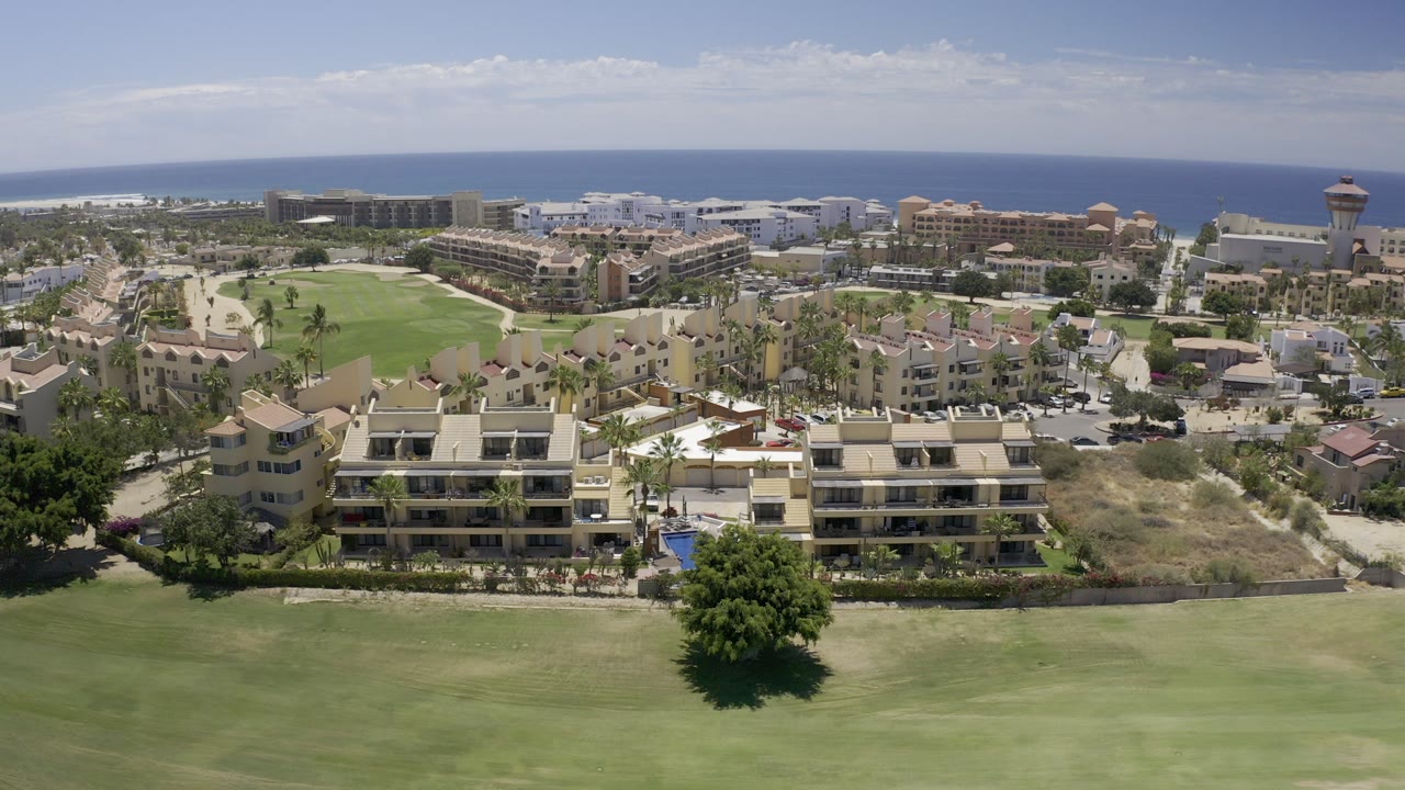 High aerial view in Los Cabos, Mexico, with a broad green field in front, beige residential buildings beyond it, the ocean on the horizon, and no visible people.