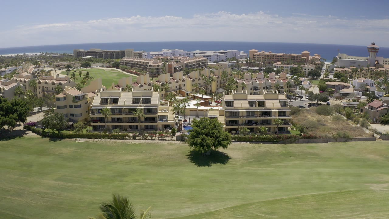 Opening aerial frame in Los Cabos shows a broad green lawn, beige multi-story building complexes, palm trees, and the ocean horizon under bright daylight, with no visible people.
