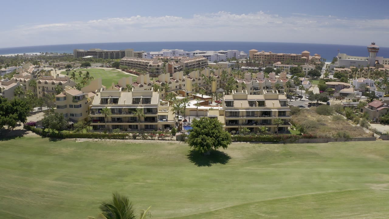 At 0:01, the Los Cabos aerial view holds on the green open space with clustered beige buildings, palms, distant rooftops, and a flat blue sea line, with no visible people.