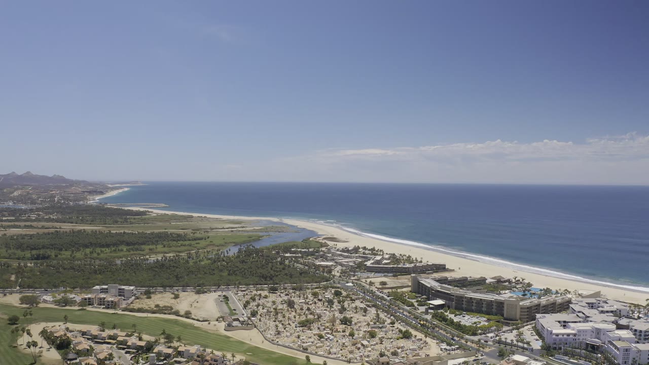 The next Los Cabos aerial frame keeps the beach and ocean horizon prominent while low coastal buildings, sandy lots, roads, and planted palms fill the lower frame, with no visible people.