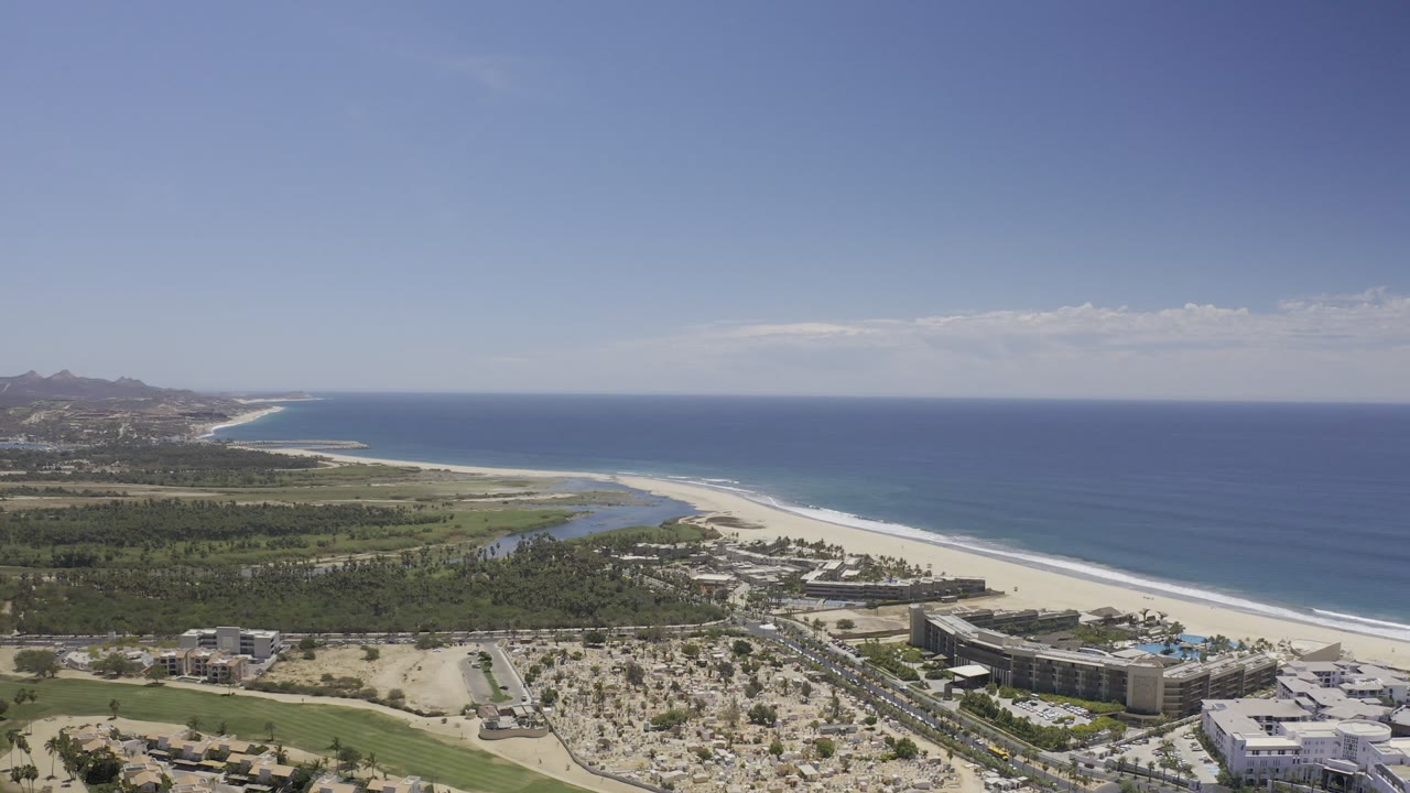 Wide aerial view over the Los Cabos coastline with white sand beach, blue ocean, green land, resort buildings, distant hills, and no visible people.