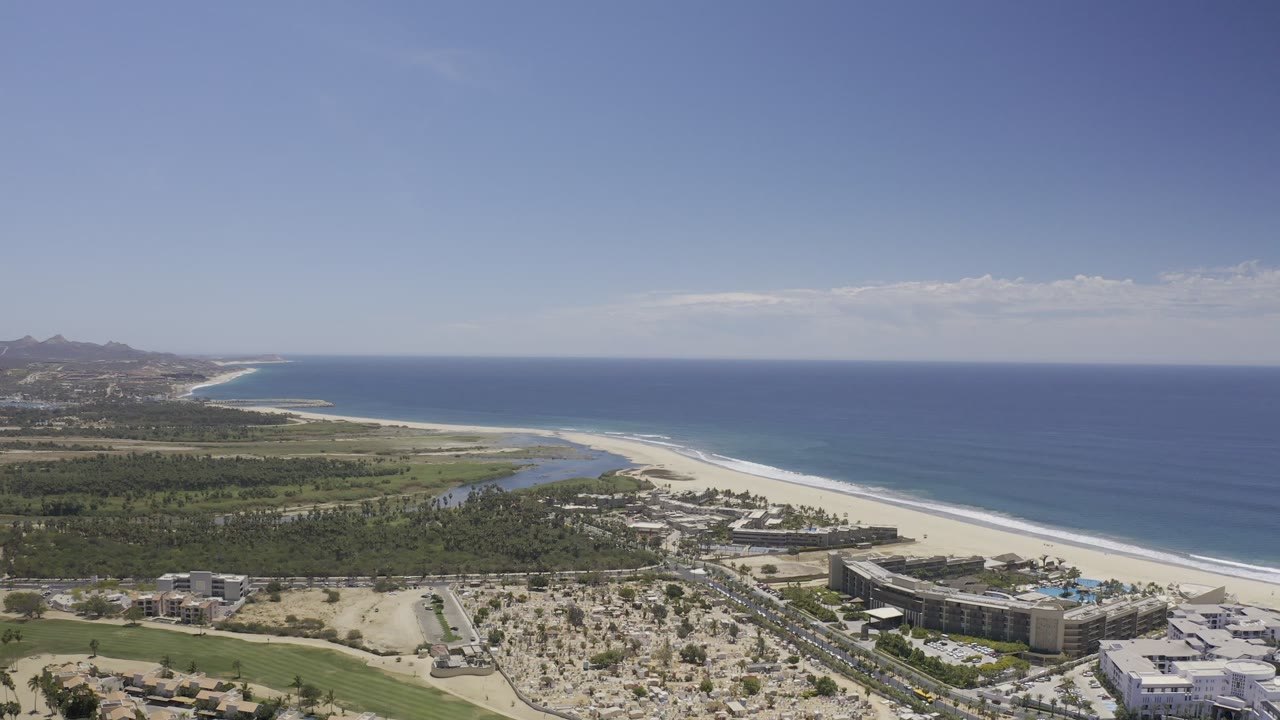 High coastal view with blue sea, sandbar-like beach, inland green areas, and buildings.