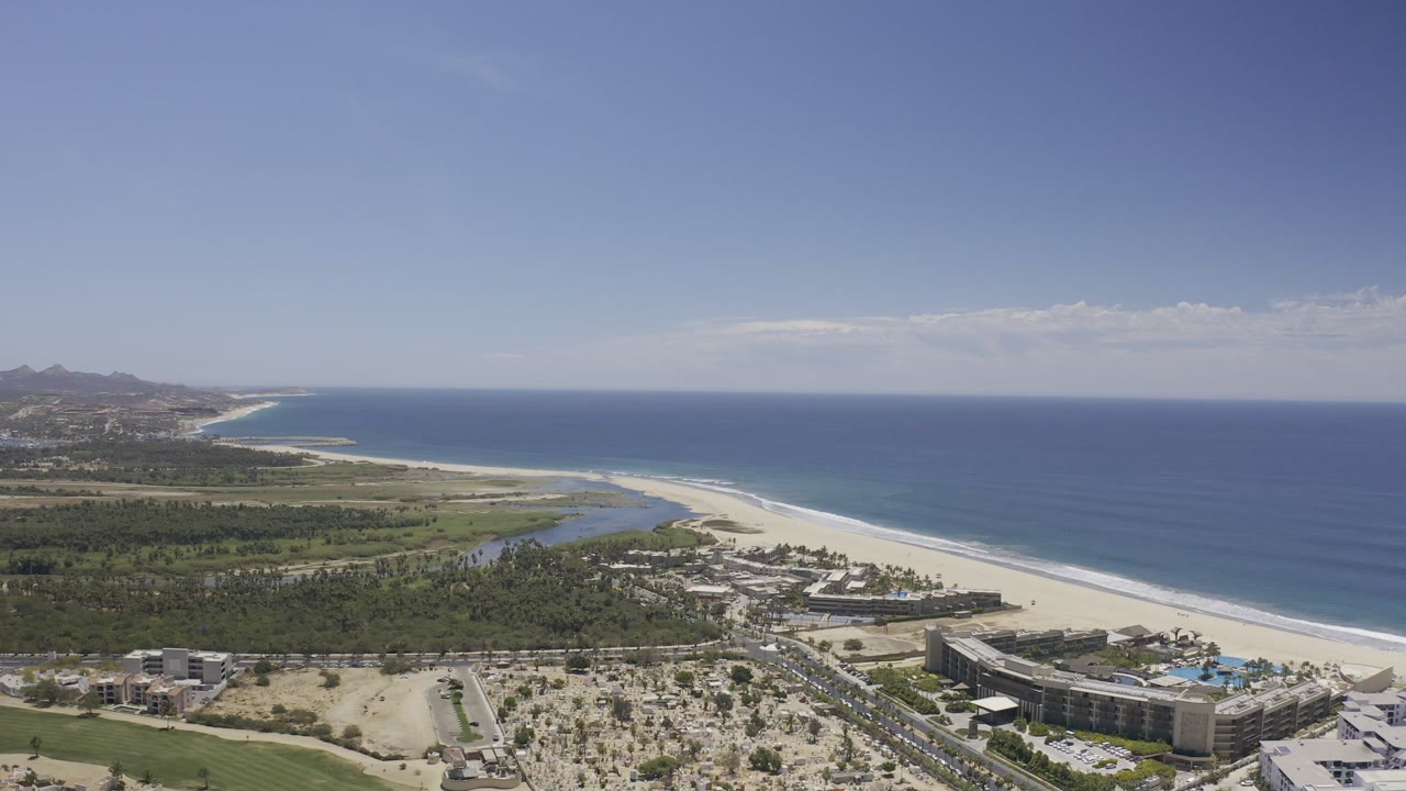 Aerial view over Los Cabos, Mexico showing blue ocean, a long white sand beach, green coastal land, roads, and multi-story resort buildings, with no visible people.