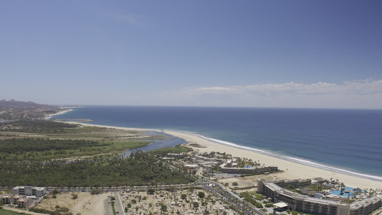 High drone view over the Los Cabos coastline with pale beach, blue sea, lagoon outlet, green inland area, roads, buildings, and no visible people.