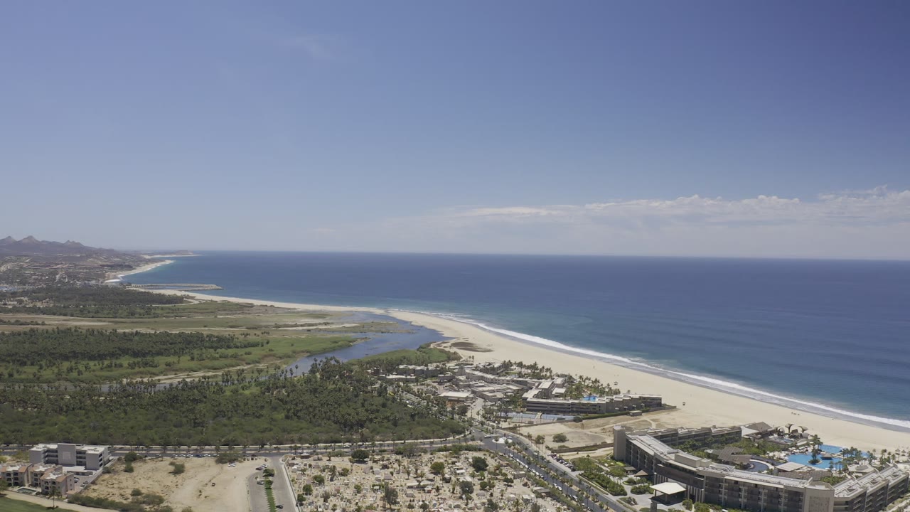 Los Cabos aerial frame showing the shoreline slightly farther along, with blue ocean to the right, beach through the center, inland greenery, low buildings, and no visible people.