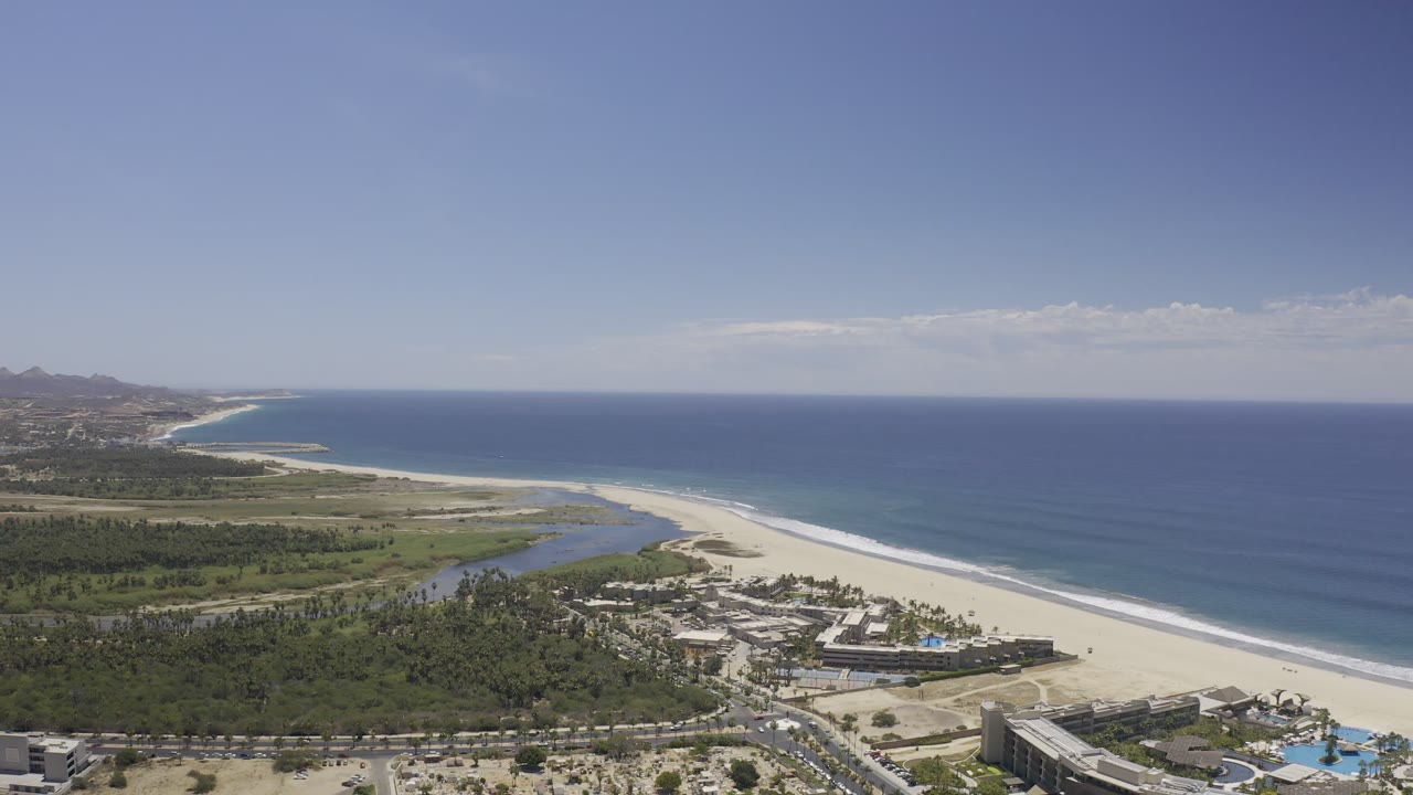Aerial frame from Los Cabos, Mexico, showing the shoreline angled across the scene, blue ocean to the right, green vegetation around a lagoon, foreground roads, and no visible people.