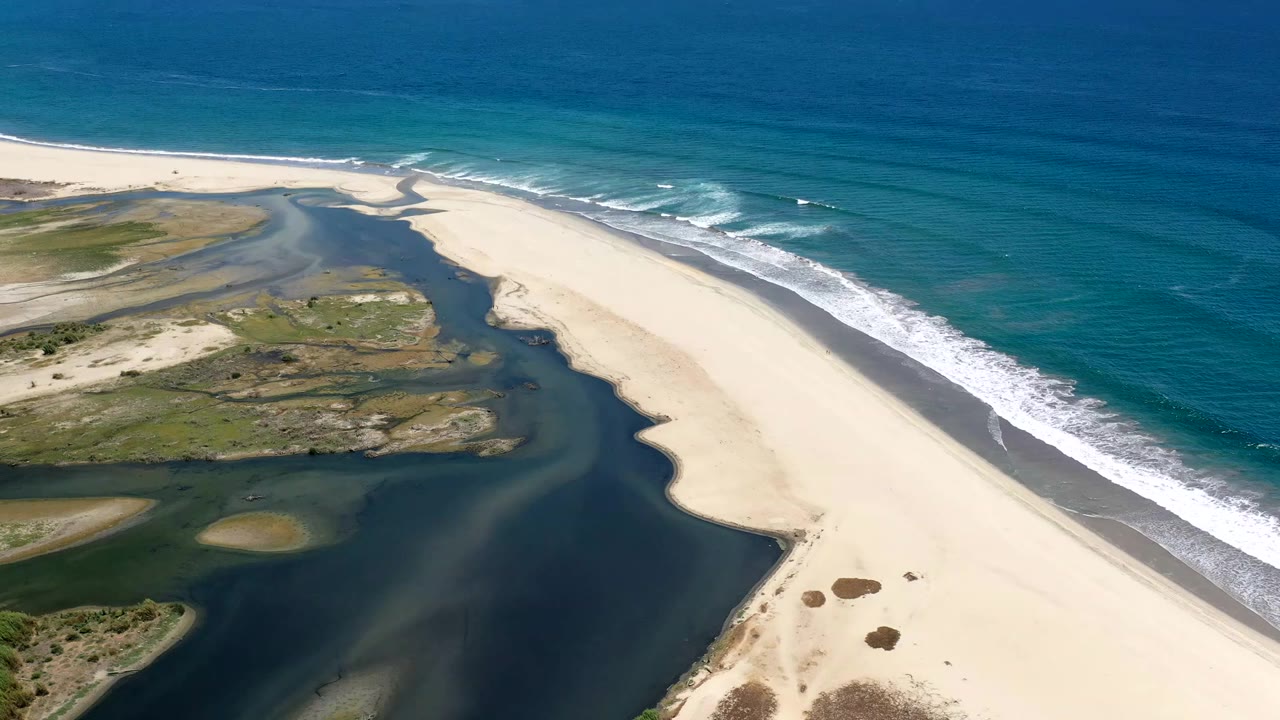 Aerial frame from Los Cabos, Mexico showing a wide pale sand beach separating dark lagoon water from blue ocean surf, with no visible people.