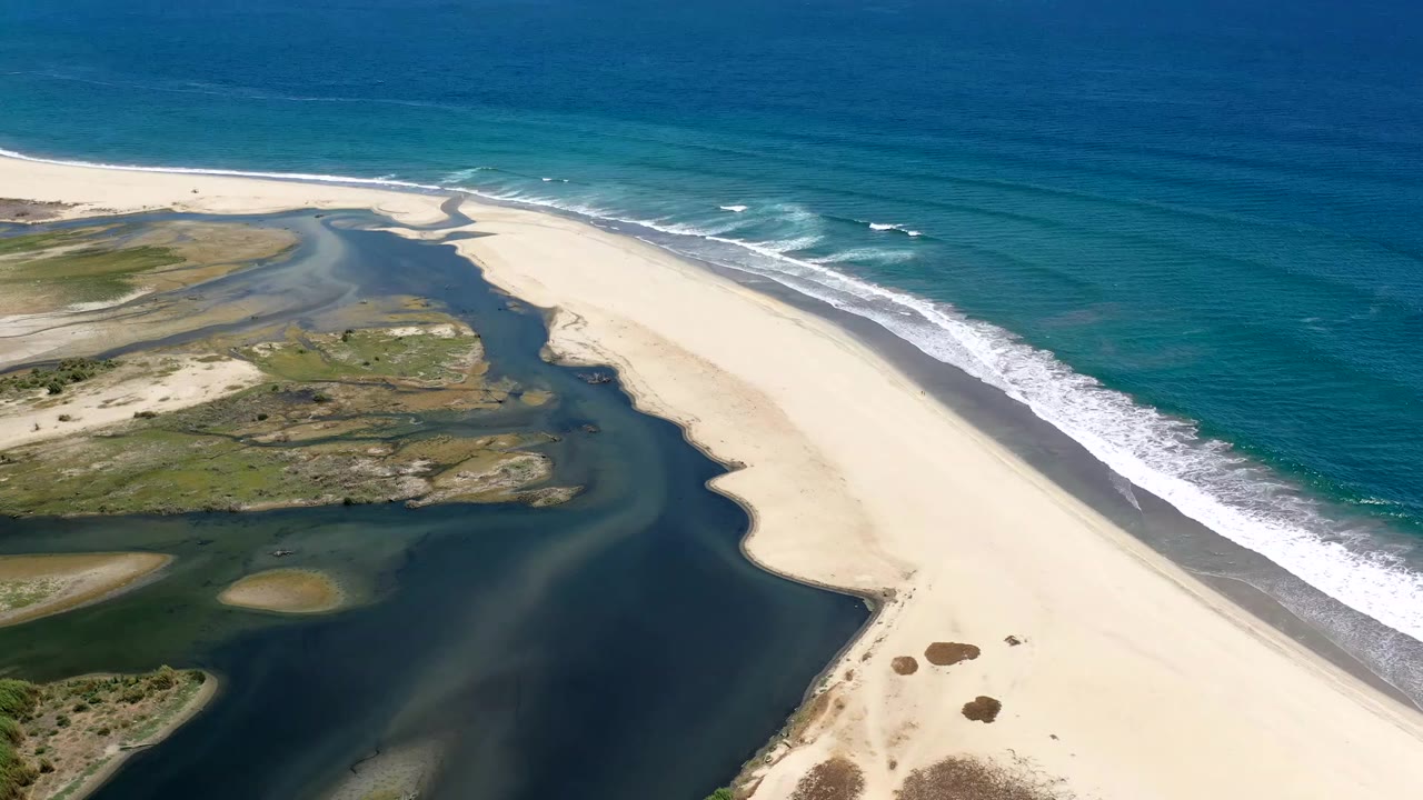 Aerial view over Los Cabos coastline with lagoon channels on the left, pale sand in the center, and turquoise ocean waves on the right, with no visible people.